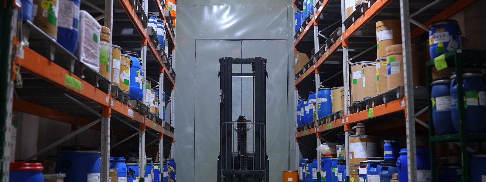 Aisle of orange used pallet racking loaded with drums and a forklift in a warehouse